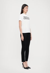Balta marškinėliai su juodu užrašu "VERSACE JEANS COUTURE", juodos laisvos kelnės ir juodi aštriais smailiais bateliai, stovintys prieš paprastą foną.