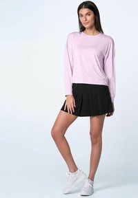 Donna in piedi con una camicia a maniche lunghe rosa chiaro, una gonna nera plissettata e delle sneaker bianche su uno sfondo chiaro uniforme.