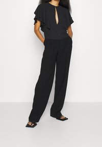 Donna che indossa pantaloni larghi neri, top nero con maniche a ruches e spacco anteriore, mani nelle tasche e sandali platform neri con punta quadrata.
