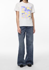 Bílá bavlněná tričko s geometrickým vzorem v purpurové a žluté barvě, spárované s vysokým pasem a širokými nohavicemi z tmavého denimu.