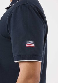 Vue de profil d'une personne portant un polo bleu marine à manches courtes avec une bordure blanche et un écusson "Seaside Nautical Legacy" sur la manche.