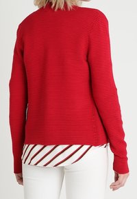 Roter strukturierten Pullover mit langen Ärmeln, gestaltet mit einem gerippten Muster. Getragen über einem weißen Hemd mit roten und schwarzen Streifen, kombiniert mit weißen Hosen.