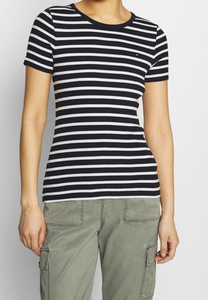 Henkilö, jolla on mustavalkoraidallinen lyhythihainen paita ja vihreät cargo-housut, seisoo yksinkertaisen vaalean taustan edessä.