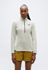 Γυναίκα που φοράει εκρού φλις με φερμουάρ μέχρι το στήθος και μουσταρδί cargo σορτς, στέκεται μπροστά σε απλό ανοιχτό γκρι φόντο.