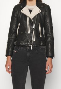 Chaqueta de motocicleta de cuero negro con un cuello beige claro, cremalleras asimétricas y bolsillos frontales. Lleva puesta con jeans oscuros.