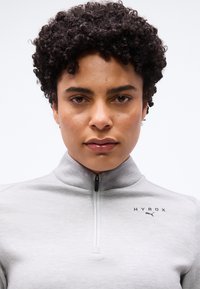 Άτομο με κοντά σγουρά μαλλιά που φοράει ένα ανοιχτό γκρι ζακέτα με φερμουάρ, με τα λογότυπα "HYROX" και Puma στο στήθος, κοιτάζοντας μπροστά.