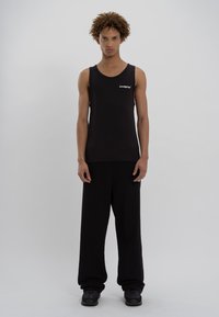 Sort ærmeløs tanktop med ribbet tekstur, der har et lille logo. Sammen med brede sorte bukser og sort fodtøj. Enkel, minimalistisk design.