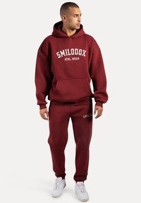 Conjunto de sudadera con capucha y pantalones de chándal burdeos, hecho de tela sólida, con un bolsillo frontal y "SMILODOX ATHL. WEAR" impreso en blanco. Zapatillas blancas.
