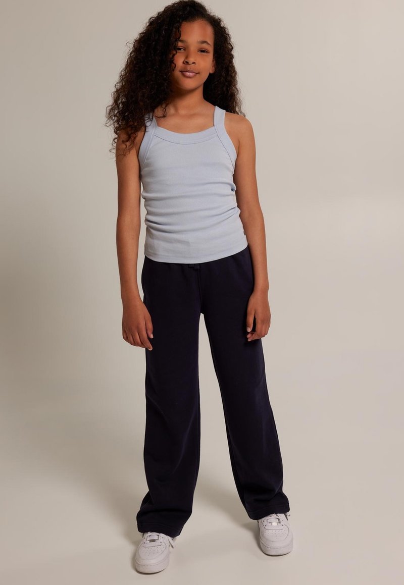Niña de pie con el pelo largo y rizado, vistiendo una camiseta sin mangas azul claro, pantalones negros y zapatillas blancas, sobre un fondo liso.