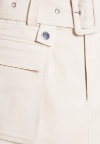 Detailní záběr na béžové látkové kalhoty s velkou kapsou, kovovým nýtem, poutkem na opasek a odpovídajícím béžovým opaskem s obdélníkovou sponou.