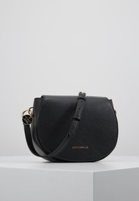 Borsa a tracolla in pelle nera con texture, flap curvo, tracolla regolabile, accessori in tonalità oro e marchio "COCCINELLE" sul davanti.