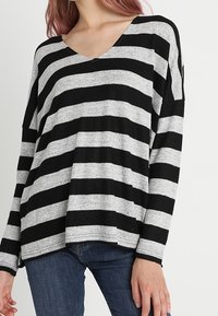 Schwarz und grau gestreiftes Langarmoberteil mit V-Ausschnitt, lockerer Passform und weichem Material, kombiniert mit blauen Jeans.