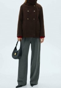 Cappotto doppiopetto in lana marrone con bottoni dorati, collo alto e tasche laterali, abbinato a pantaloni grigi a gamba larga e a una borsa nera.