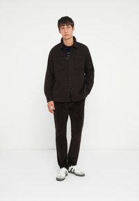 Black shirt jacket with button closure, two chest pockets, paired with matching black pants, white sneakers with black stripes.