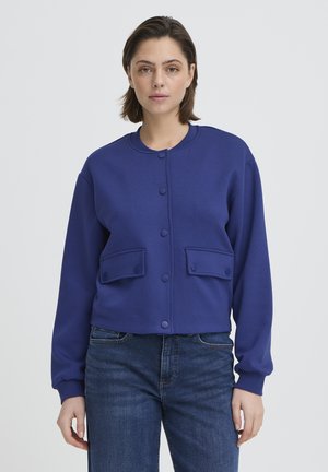 Femme portant un cardigan bleu à manches longues boutonné avec poches avant et un jean bleu, debout devant un fond clair uni.