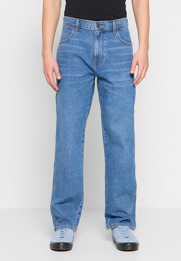 Niebieskie dżinsy o luźnym kroju, z pięcioma kieszeniami i akcentami w postaci przeszyć. Gładka tekstura i prosty krój nogawki, zestawione z jasnym obuwiem.