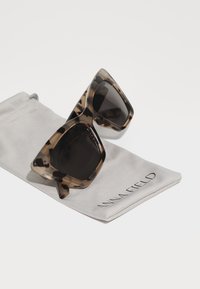 Black and brown tortoiseshell sunglasses with angular frames, resting on a gray soft pouch featuring "ANNA FIELD" in black lettering.
