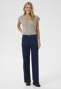 Jasnoszara koszulka V-neck w połączeniu z ciemnoniebieskimi spodniami w szerokie nogawki, które mają przednie i tylne kieszenie. Modelka stoi na jednolitym tle.
