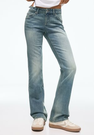 Person trägt hellblaue ausgewaschene Jeans mit geradem Bein und weiße sowie beige Sneaker und steht mit den Händen in der Taille.