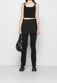 Schwarzes geripptes Crop-Top mit verstellbaren Trägern, kombiniert mit einem schwarzen Wickelrock und enganliegenden Leggings. Das Model hält eine geknotete schwarze Handtasche.