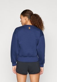 Μπλε ναυτικού φούτερ φτιαγμένο από μαλακό υφασμά, με ριχτό στρίφωμα και μανίκια, και μια μικρή ετικέτα λογότυπου στο πίσω μέρος. Φοριέται με μαύρες βερμούδες.