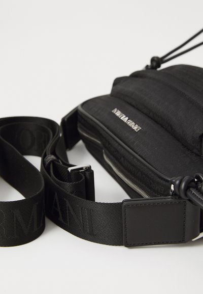 Geantă crossbody neagră cu design căptușit, având închidere cu fermoar și model logo embosat. Curea reglabilă cu detalii logo și accesorii din metal.