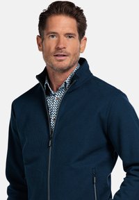 Navyblauwe ritsjack met een gestructureerd oppervlak en hoge kraag, gedragen over een gestreept wit shirt met kleine blauwe en zwarte accenten.