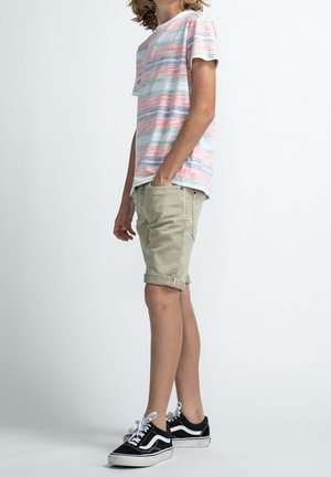 T-shirt a righe in rosa, verde e blu. Pantaloni corti beige arrotolati. Sneakers nere con dettagli bianchi. Posizione in piedi su uno sfondo neutro.