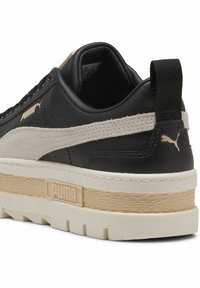 Sneaker nera in pelle con dettagli in suede beige, suola bianca spessa e intersuola testurizzata. Presenta un logo sul tallone e cuciture decorative.
