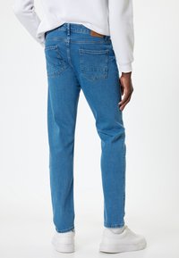 Pantalones vaqueros de denim azul con un corte ajustado, dos bolsillos traseros y una etiqueta de logo de cuero marrón en la cintura, combinados con zapatillas blancas.