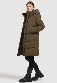 Olivgrüne Pufferjacke mit Kapuze, mit einem Reißverschluss an der Vorderseite, seitlichen Taschen und gepolsterter Textur. Kombiniert mit schwarzen Schnürstiefeln.