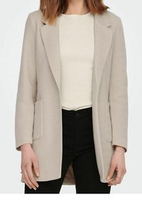 Beige blazer s rybí kostrou se dvěma předními kapsami, propracovaný design, strukturované klopy, nošený přes světlobéžový top a černé kalhoty.