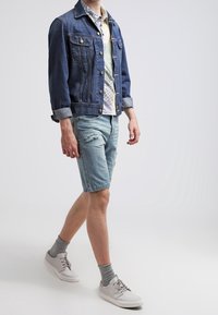 Veste en jean avec boutons sur le devant et poches poitrine, associée à une chemise à manches courtes à motifs, un short délavé clair déchiré et des baskets blanches.