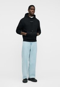 Νεαρός άνδρας που στέκεται με τα χέρια στις τσέπες, φοράει μαύρο φούτερ με κουκούλα, ανοιχτό μπλε τζιν με φαρδύ πόδι και μαύρα αθλητικά, μπροστά σε απλό λευκό φόντο.