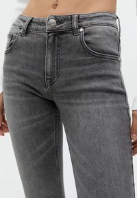 Läheltä kuvattu henkilö, joka on pukeutunut korkeavyötäröisiin, haalistuneisiin mustiin skinny-farkkuihin, joissa on nappi- ja vetoketjukiinnitys sekä taskut kummallakin puolella.