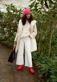 Gilet matelassé beige par-dessus un pull crème, jean large blanc, sabots rouges, bonnet rose, tenant un sac noir avec des accents rouges et blancs, entouré de verdure.
