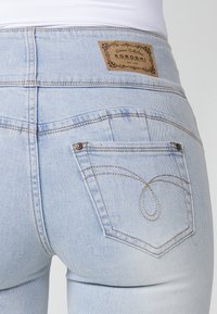 Jeans de mezclilla azul claro con cintura alta, que presentan una etiqueta de cuero con la palabra "KOROSHI" y costuras decorativas en los bolsillos traseros.