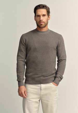 Grauer gestrickter Pullover mit Rundhalsausschnitt und langen Ärmeln, der ein strukturiertes Muster aufweist. Kombiniert mit hellfarbigen Hosen.