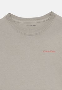 T-shirt en coton gris à col ras du cou, arborant un petit logo rouge "Calvin Klein" sur le côté gauche de la poitrine. Texture lisse.