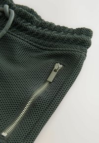 Grå mesh-jogger med elastisk midjeband, med en sidoficka med dragkedja och texturerat tyg. Detaljer inkluderar metallic dragkedja.
