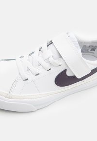 Sapatilha branca com um cabedal de couro liso, fecho por atacadores e tiras com fecho de gancho, e o logotipo da Nike em roxo. Sola texturizada.