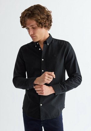 Mężczyzna z kręconymi włosami zapinający mankiet czarnej koszuli z długim rękawem i białymi guzikami, stojący na tle jasnego, jednolitego tła.