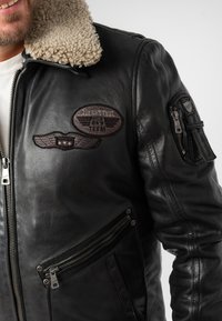 Schwarze Lederjacke mit Schafslederkragen, mehreren Reißverschlusstaschen und gestickten Patches auf der Brust und dem Ärmel.