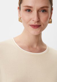 Beżowy dzianinowy top z okrągłym dekoltem i kontrastowymi białymi wykończeniami. Model nosi złote kolczyki w kształcie kół. Gładka tekstura, dopasowany fason.