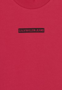 Červená tkanina s malou černou obdélníkovou nálepkou umístěnou uprostřed, na které je velkými písmeny napsáno "CALVIN KLEIN JEANS".
