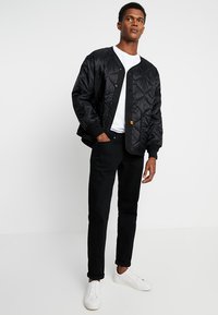 Black quilted bomber jacket with ribbed cuffs, button closure, and an orange logo tag. Paired with black pants and white sneakers.