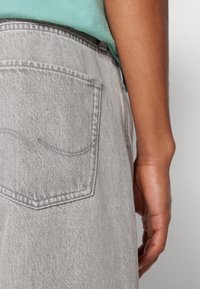 Detailní záběr ruky osoby odpočívající vedle světle šedé denimové kapsy a světle zelené košile zasunuté dovnitř.