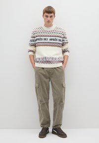 Gestrickter Pullover in Creme mit roten und marineblauen geometrischen Mustern. Olivgrüne Cord-Cargohosen mit Seitentaschen. Braune Sneaker mit dunklen Akzenten.