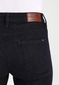 Tummansiniset farkut, joissa on nahkainen logo-ompelus vyötäröllä, sekä takatasku hienovaraisilla ompelu yksityiskohdilla ja pienellä lipputingalla.