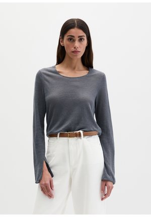 Vrouw met lang donker haar die een grijs longsleeve shirt met splitten en witte hoge taille broek met een bruine riem draagt, staand.